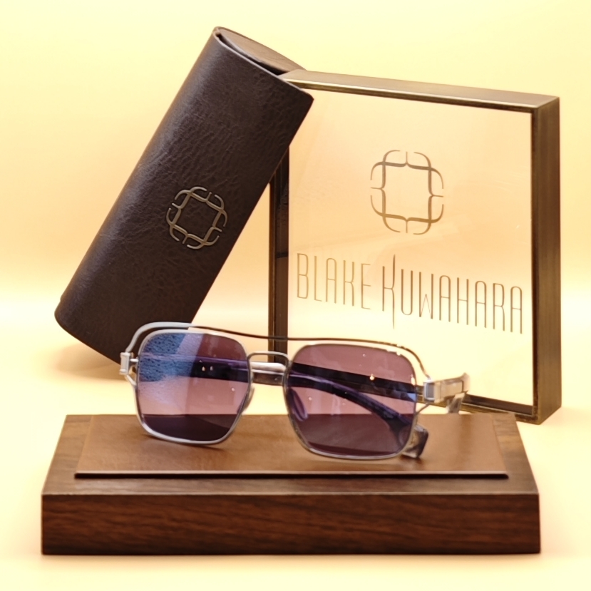 Blake Kuwahara's Hasting metal sunglasses with purple-tinted lenses, an aviator-style double bridge, and clear acetate accents, displayed on a wooden stand next to a dark brown case and branded box.