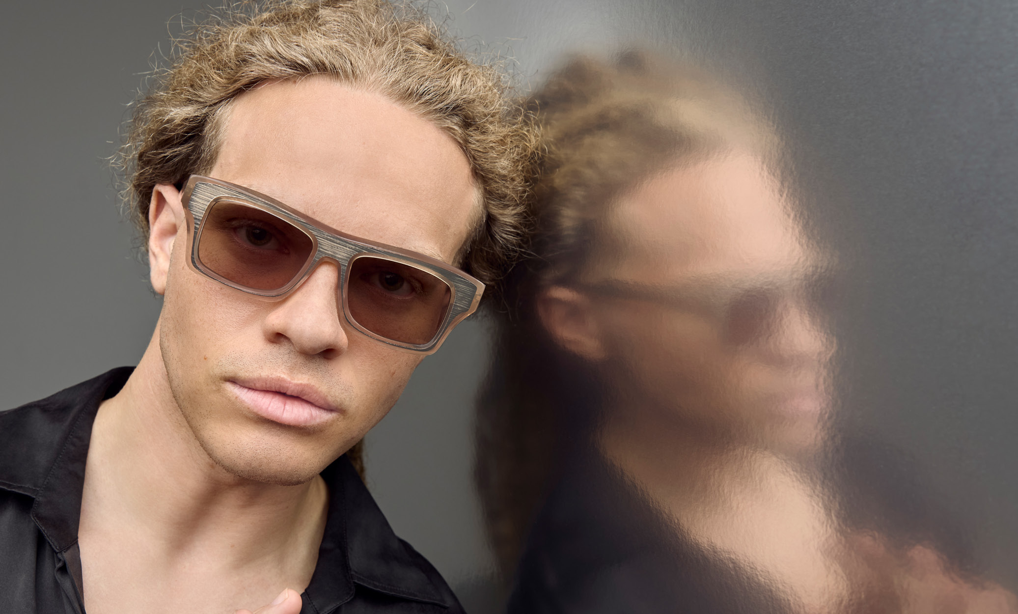 Close-up of a model wearing Blake Kuwahara thick-rimmed tan sunglasses with textured gray upper frames, leaning against a reflective metallic wall.