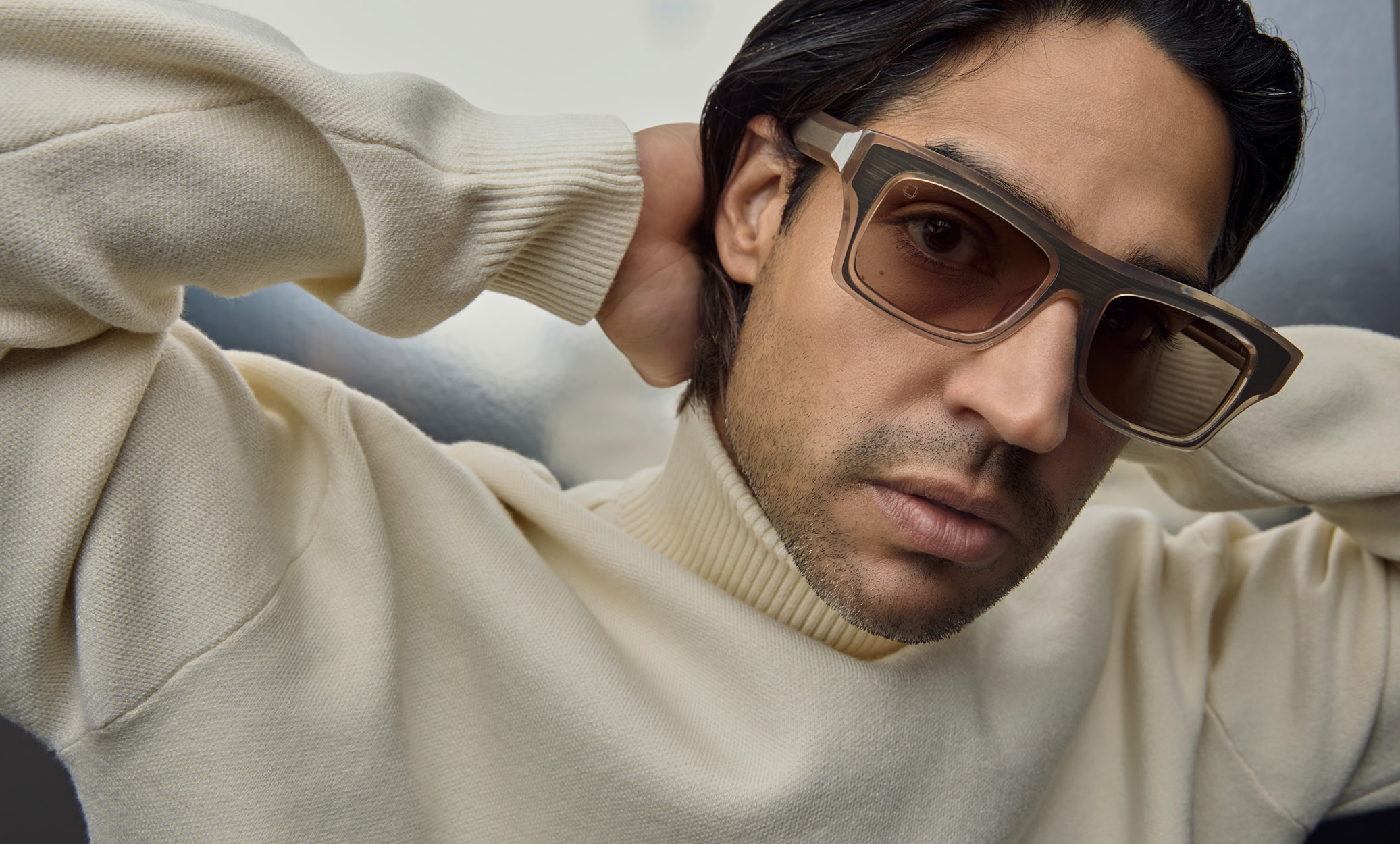 Close-up of a model wearing Blake Kuwahara thick-rimmed tan sunglasses with textured gray upper frames, leaning against a reflective metallic wall.